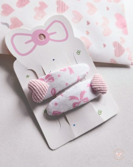 Set of two handmade hair-clips made of white fabric with pink bows and with pink heart-shaped details added.