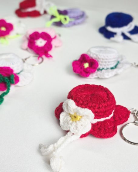 Colourful crocheted keychains in various shapes, featuring hats with  flowers and decorative details.