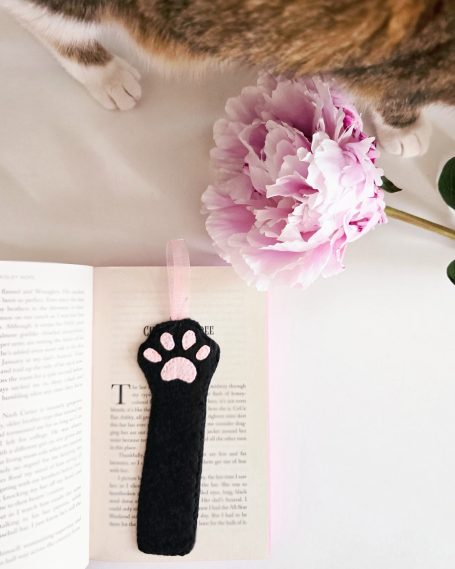 A black cat paw bookmark on an open book next to a pink peony and a cat's paws.