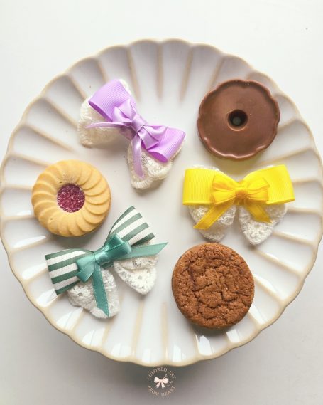 Handmade pastel mini bow set inspired by candy colours (sage green, lavander and yellow) displayed alongside cookies on a plate.