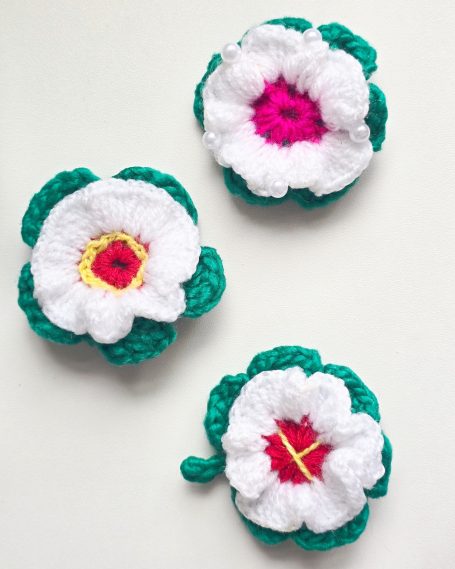 Three crocheted flowers with white petals and green leaves, featuring pink and yellow centres.