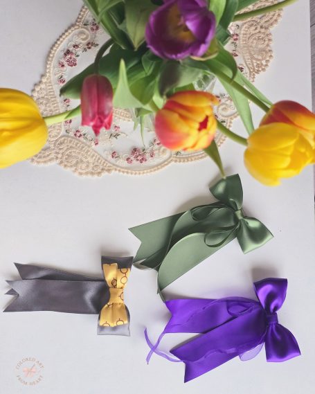 Handmade decorative bows set of three, in purple, gold and green next to vibrant tulips.