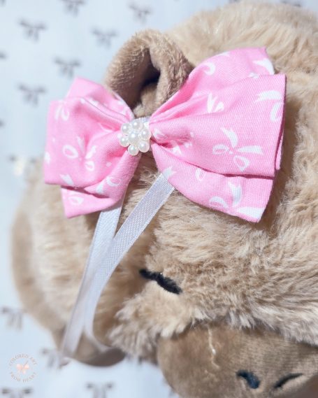 Handmade hair bow made of pink fabric with white bows, featuring a small flower made of tiny pearls in the centre.