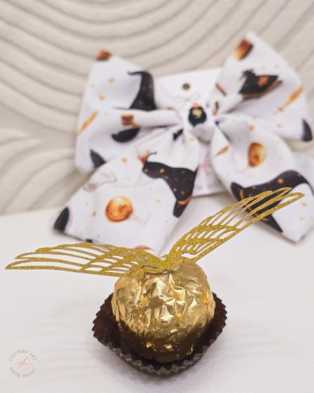 White bow with Harry Potter-themed patterns, accompanied by a gold foil chocolate with golden snitch wings.