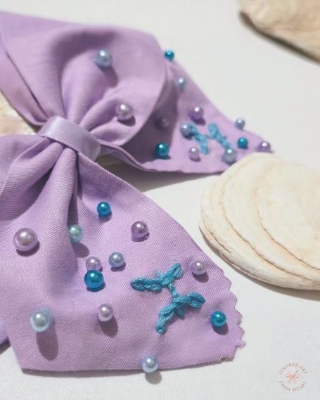 Soft lilac fabric bow with hand-embroidered Pisces symbols and tiny pearls scattered like drops of water.