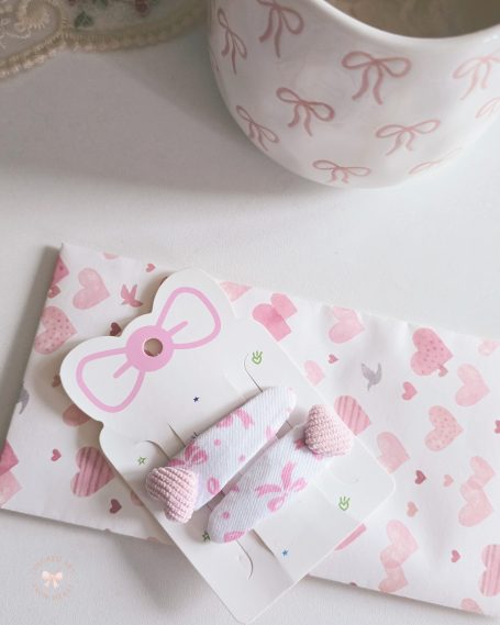 Set of two handmade hair-clips made of white fabric with pink bows and with pink heart-shaped details added.