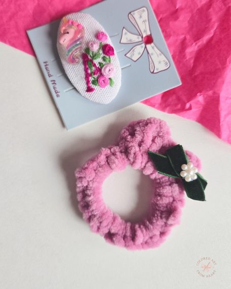 Pink scrunchie with a decorative bow paired with a floral embroidered hair clip on a patterned card.