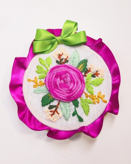 Embroidered floral design featuring a pink rose and green leaves, adorned with a ribbon.