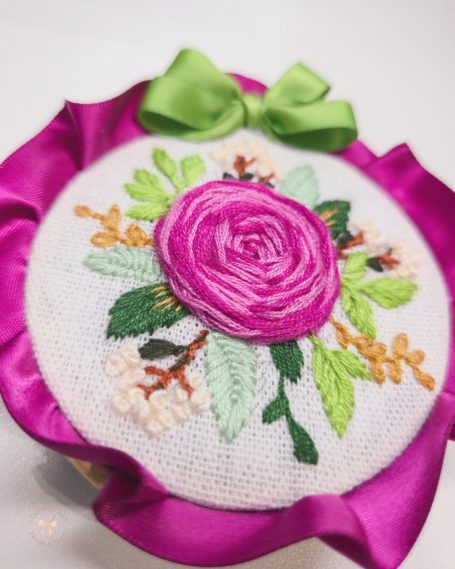 Embroidered floral design featuring a pink rose and green leaves, adorned with a ribbon.