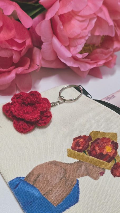 A decorative pouch featuring a painted woman in profile wearing a hat with peonies. The pouch has a crocheted peony keychain
