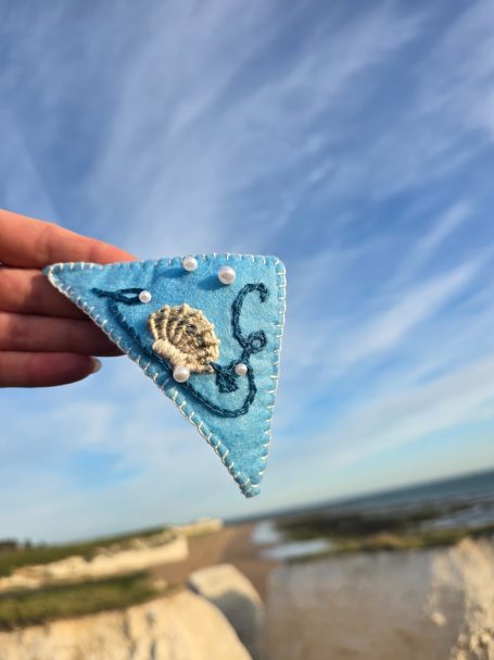 Blue felt corner bookmark with an embroidered shell and decorative beads on a beach backdrop.