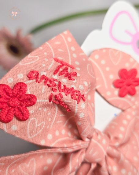 Coral pink fabric bow with red embroidered text "The answer is love", paired with a matching hair clip.