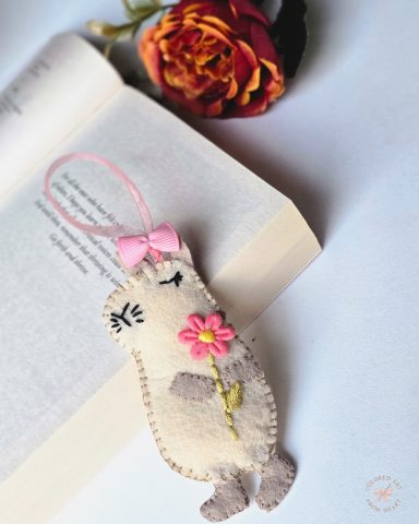 Capybara - Shaped Bookmark A handmade felt bookmark shaped like a cute capybara holding a flower, with a pink bow.