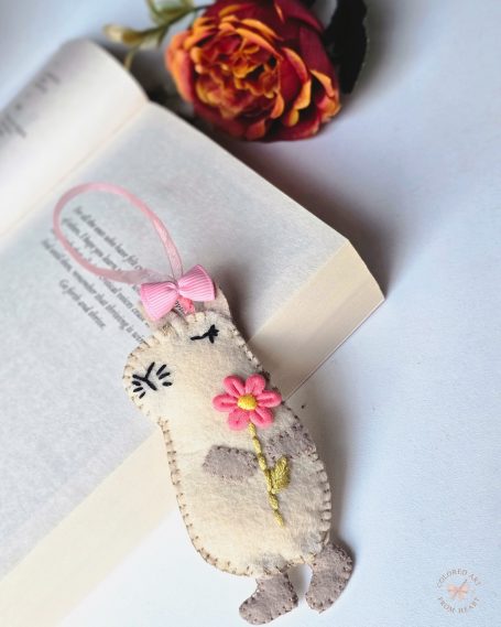 A handmade felt bookmark shaped like a cute capybara holding a flower, with a pink bow.