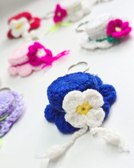 Colourful crocheted keychains in various shapes, featuring hats with  flowers and decorative details.
