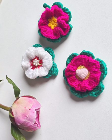 Three crocheted flowers with white petals and green leaves, featuring pink and yellow centres.