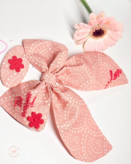 Coral pink fabric bow with red embroidered text "The answer is love", paired with a matching hair clip.