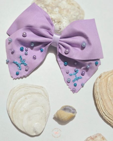 Soft lilac fabric bow with hand-embroidered Pisces symbols and tiny pearls scattered like drops of water.