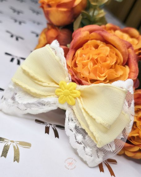 A little yellow bow with delicate white lace and a tiny, bright yellow flower in the center.