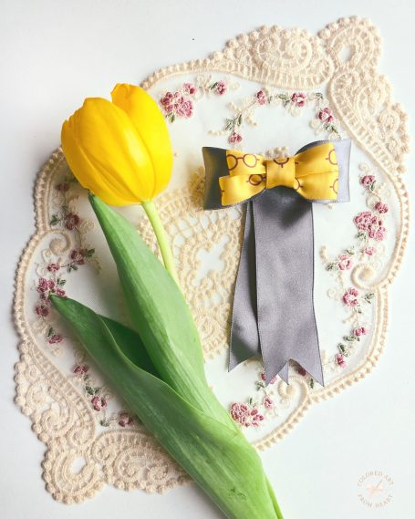 Handmade hair bow made of grey and yellow ribbons, with eyeglasses patterns, displayed with a yellow tulip.