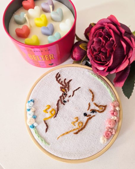 Embroidery hoop featuring deer designs with floral embellishments and a pink candle in the background.